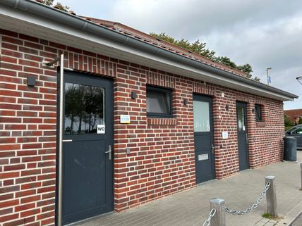 Backsteingebäude mit drei Türen zu öffentlichen Toiletten, eine davon barrierefrei. Vor dem Gebäude sind Kettenpfosten und ein Mülleimer sichtbar.