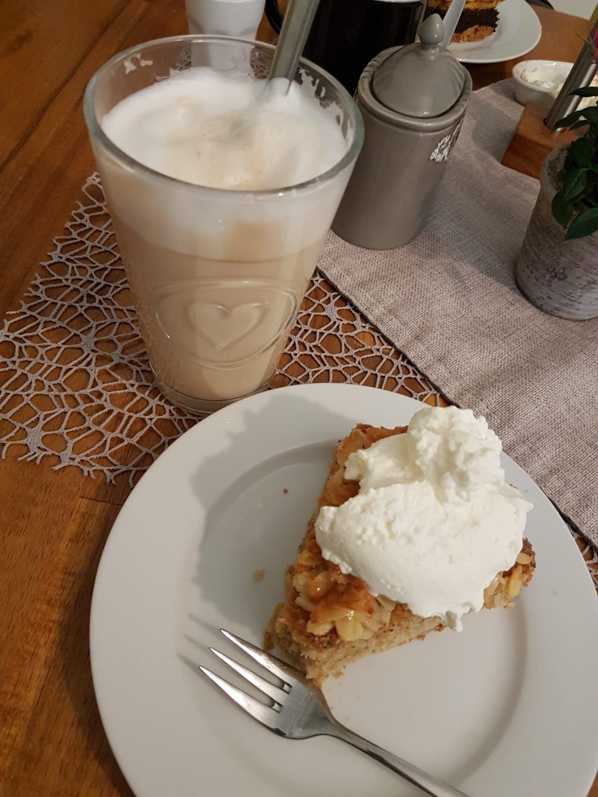 Ein Glas Latte Macchiato und ein Stück Apfelkuchen mit Sahne auf einem Teller, serviert auf einem Holztisch mit gehäkeltem Tischläufer.