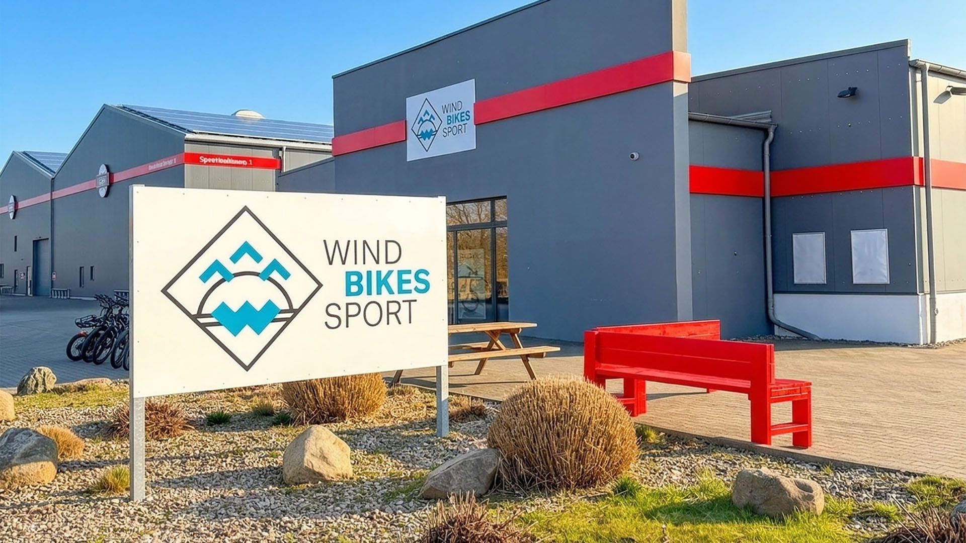 Ein modernes Gebäude mit dem Schild 'Wind Bikes Sport' und einer roten Bank im Vordergrund. Fahrräder stehen an der Seite bereit.