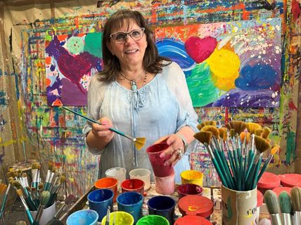Eine Frau in einem Atelier, umgeben von bunten Farben und Pinseln, lächelt vor einer farbenfrohen Leinwand.