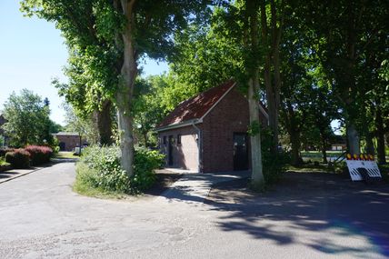 Ein kleines Backsteingebäude mit rotem Dach steht zwischen hohen Bäumen. Ein asphaltierter Weg führt daran vorbei. Im Hintergrund sind weitere Gebäude zu sehen.