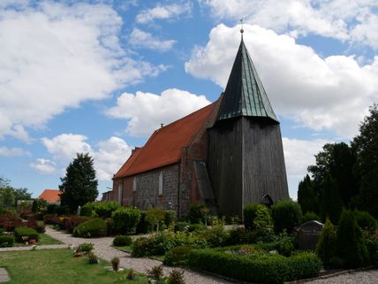 Die Kirche in Bannesdorf auf Fehmarn mit rotem Ziegeldach und hölzernem Turm, umgeben von einem gepflegten Garten unter blauem Himmel.