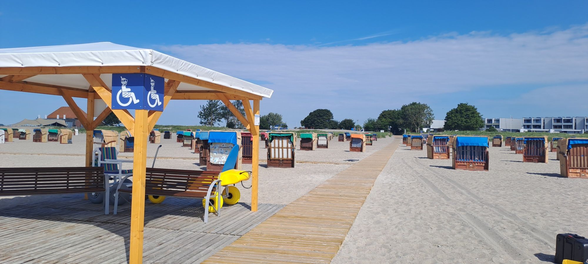 Ein barrierefreier Strandbereich mit Holzsteg, überdachtem Sitzbereich und zahlreichen Strandkörben unter blauem Himmel.