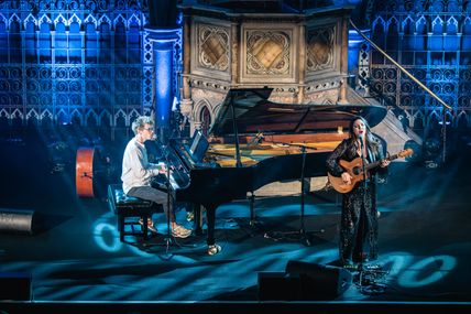 Zwei Musiker auf einer Bühne, einer am Klavier, der andere mit Gitarre. Die Bühne ist stimmungsvoll beleuchtet, im Hintergrund sind dekorative Wände zu sehen.