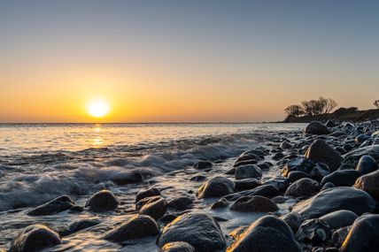 Sonnenuntergang über dem Meer mit Wellen, die auf einen steinigen Strand bei Staberdorf auf Fehmarn treffen
