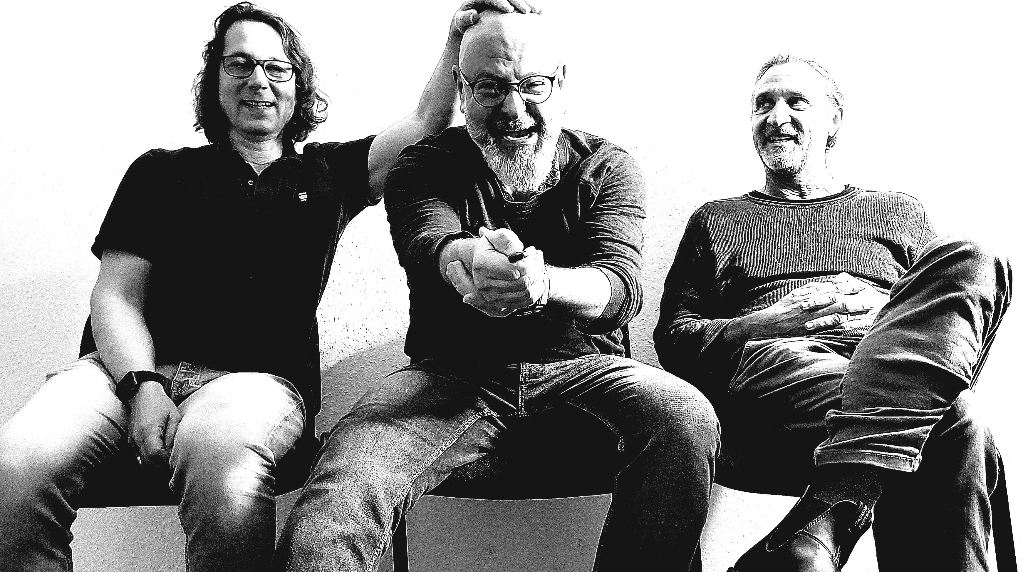 Drei Männer sitzen nebeneinander und lachen. Der mittlere Mann wird von einem der anderen am Kopf berührt. Schwarz-Weiß-Foto mit hohem Kontrast.
