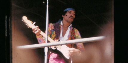 Ein Musiker mit buntem Hemd und Stirnband spielt E-Gitarre auf einer Bühne. Im Hintergrund ist ein Mikrofonständer zu sehen.