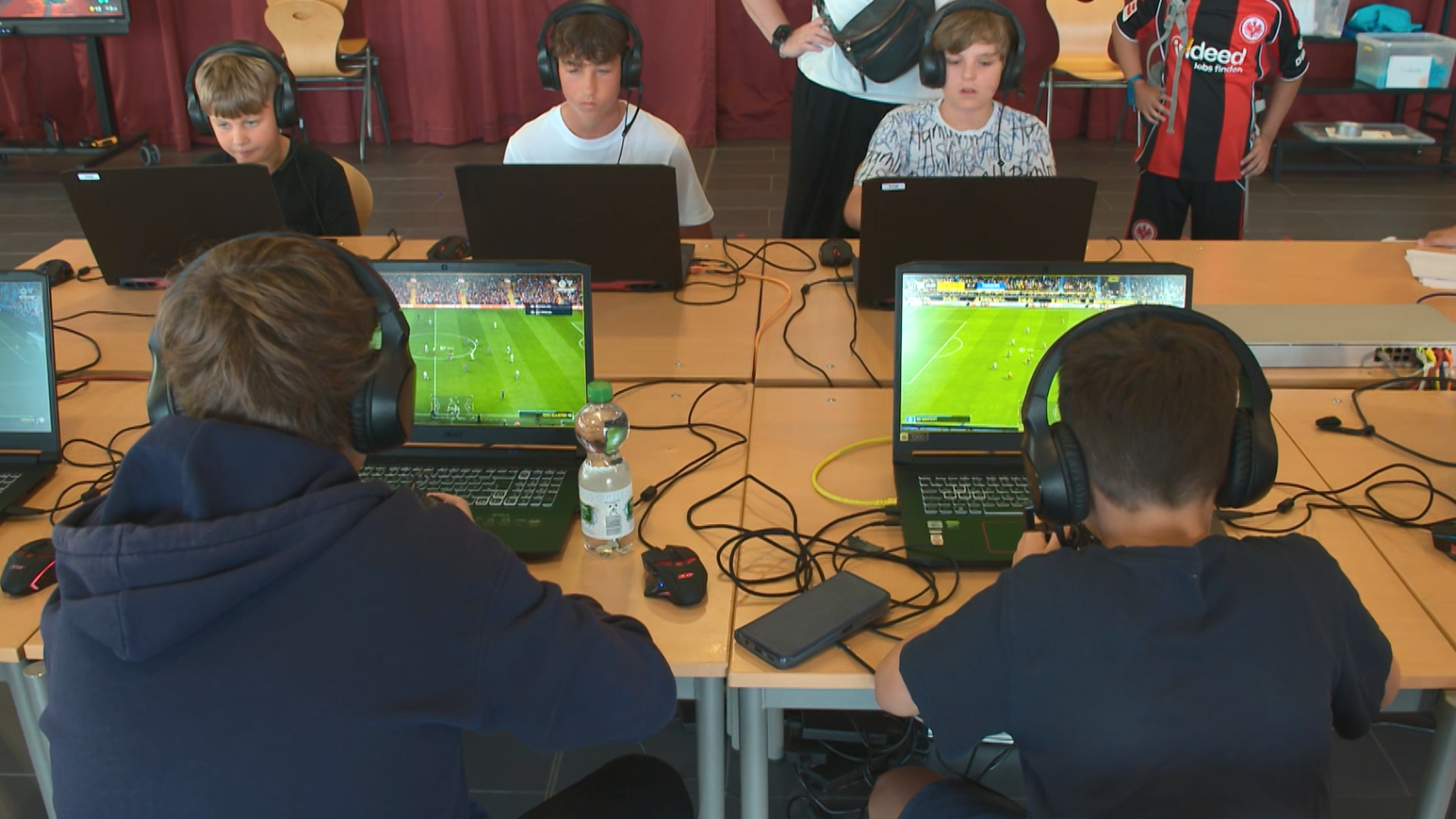 Fünf Jugendliche spielen ein Fußball-Videospiel auf Laptops. Sie tragen Kopfhörer und sind in einem Raum mit Tischen und Stühlen.