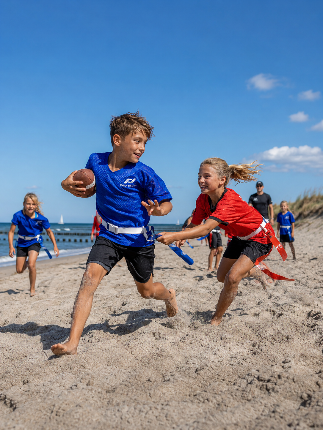 Kinder spielen Flag Football am Strand. Ein Junge in blauem Trikot läuft mit dem Ball, während ein Mädchen in rotem Trikot ihn verfolgt.