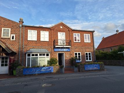 Ein Backsteingebäude mit dem Schild 'Dat lütte Fischhus' an der Fassade. Es befindet sich an einer ruhigen Straße unter blauem Himmel.