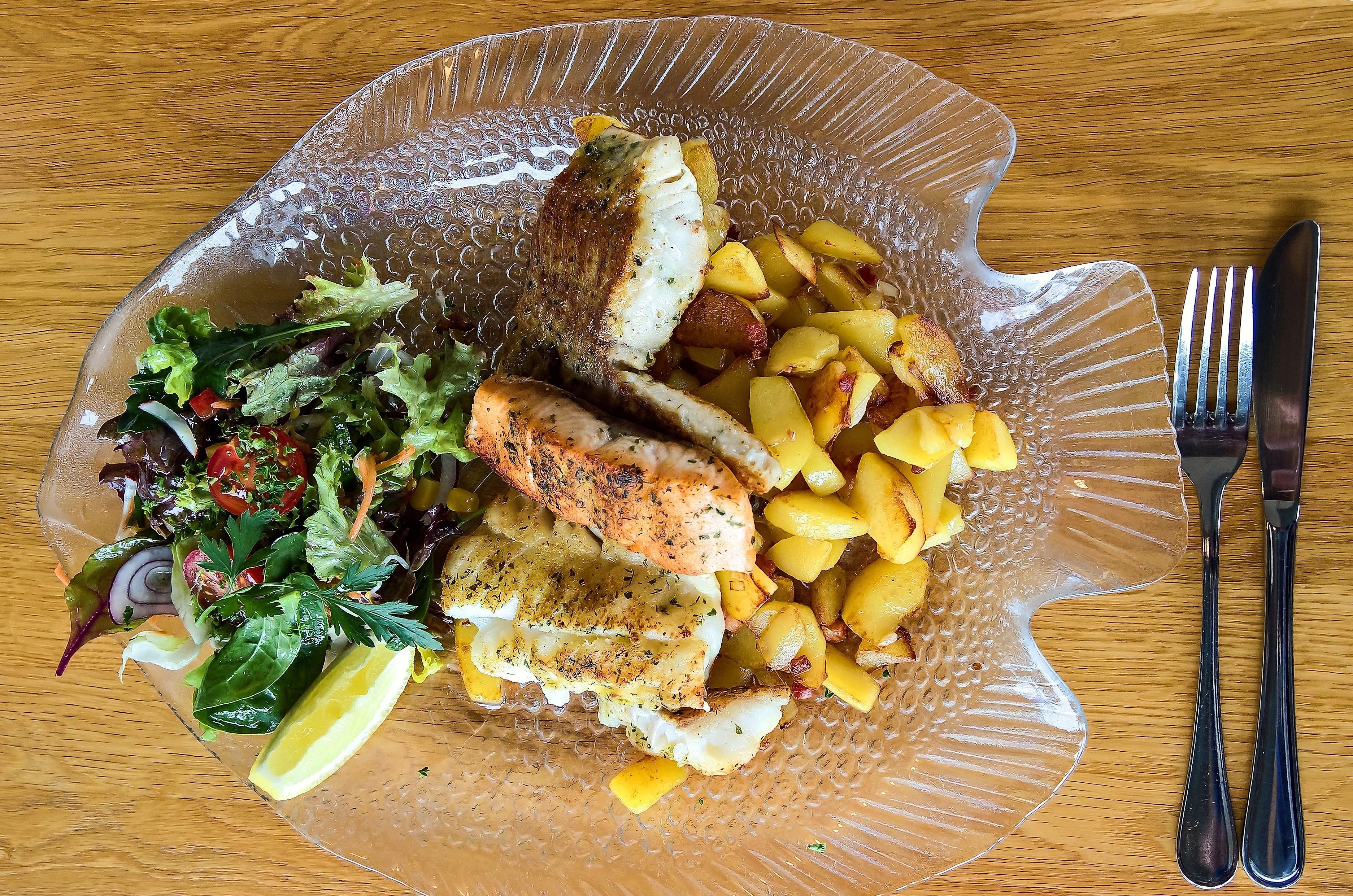 Ein Fischgericht auf einem gläsernen Teller in Fischform. Es enthält gebratenen Fisch, Bratkartoffeln und einen frischen Salat mit Zitrone.