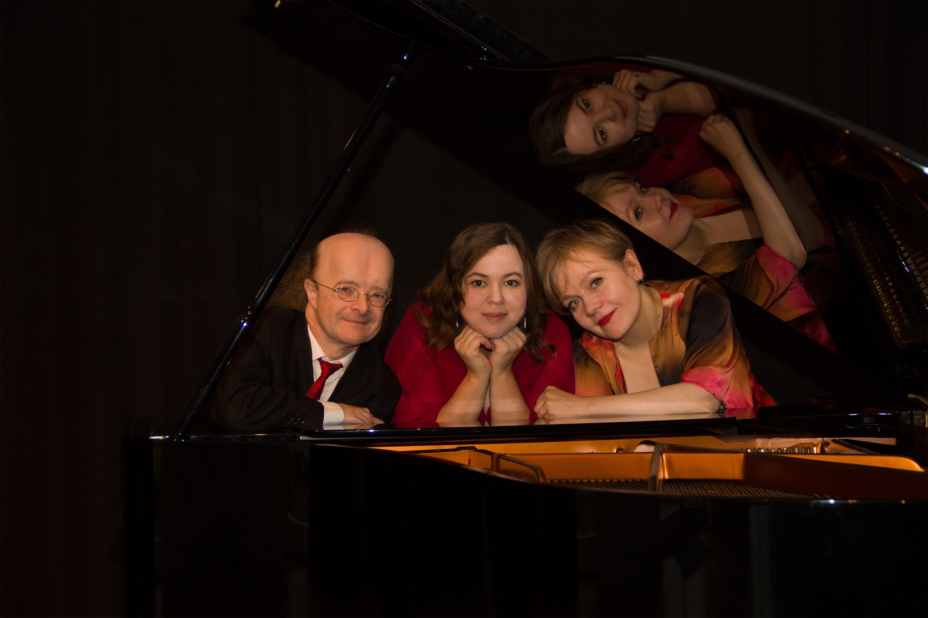 Drei Personen posieren lächelnd an einem Flügel, ihre Gesichter spiegeln sich im glänzenden Deckel wider.