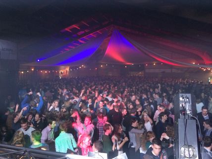 Eine große Menschenmenge tanzt in einer festlich beleuchteten Halle. Bunte Lichter und Dekorationen schaffen eine lebhafte Atmosphäre.