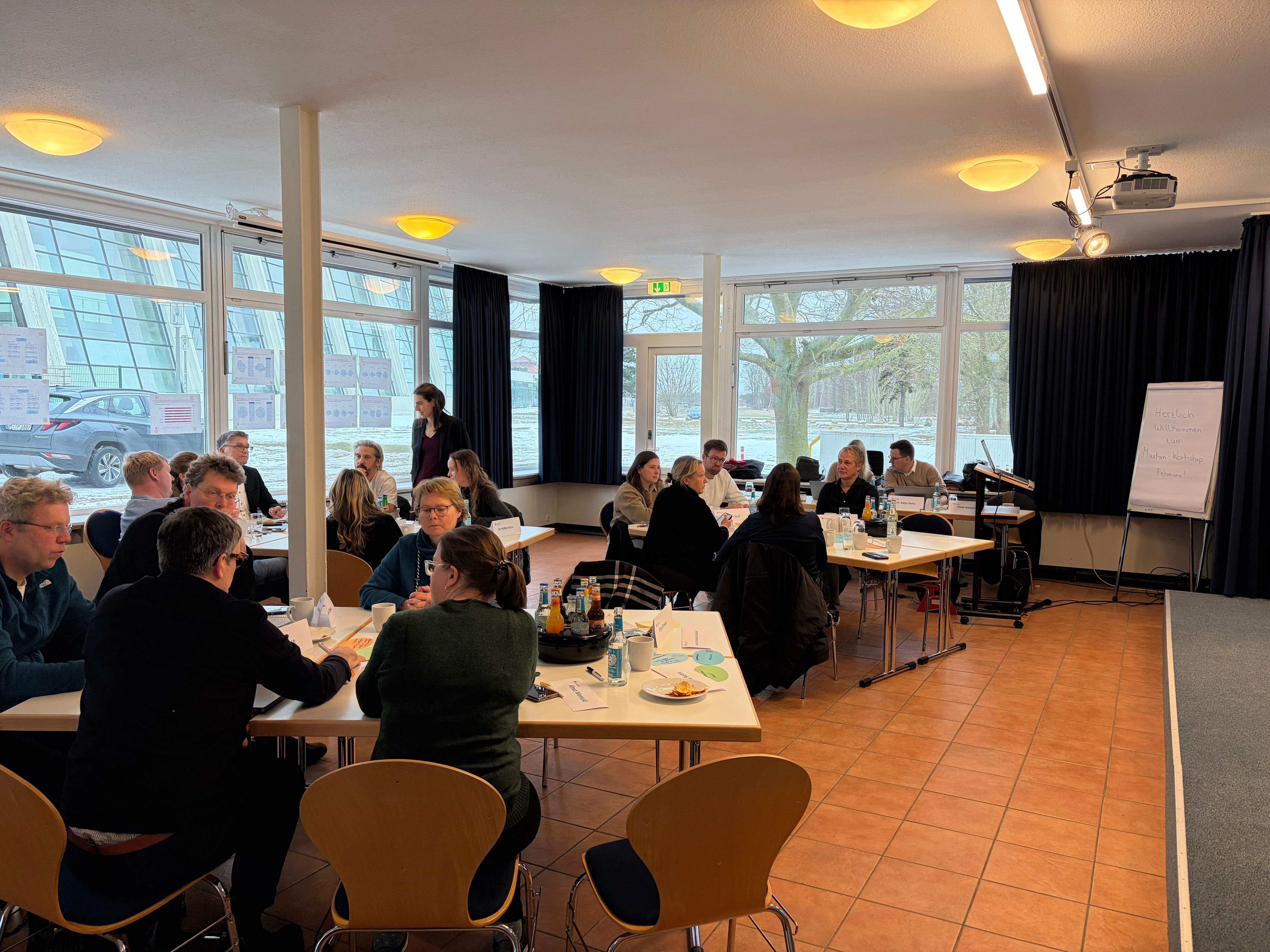 Mehrere Personen sitzen an Tischen in einem hellen Raum mit großen Fenstern und diskutieren in kleinen Gruppen.
