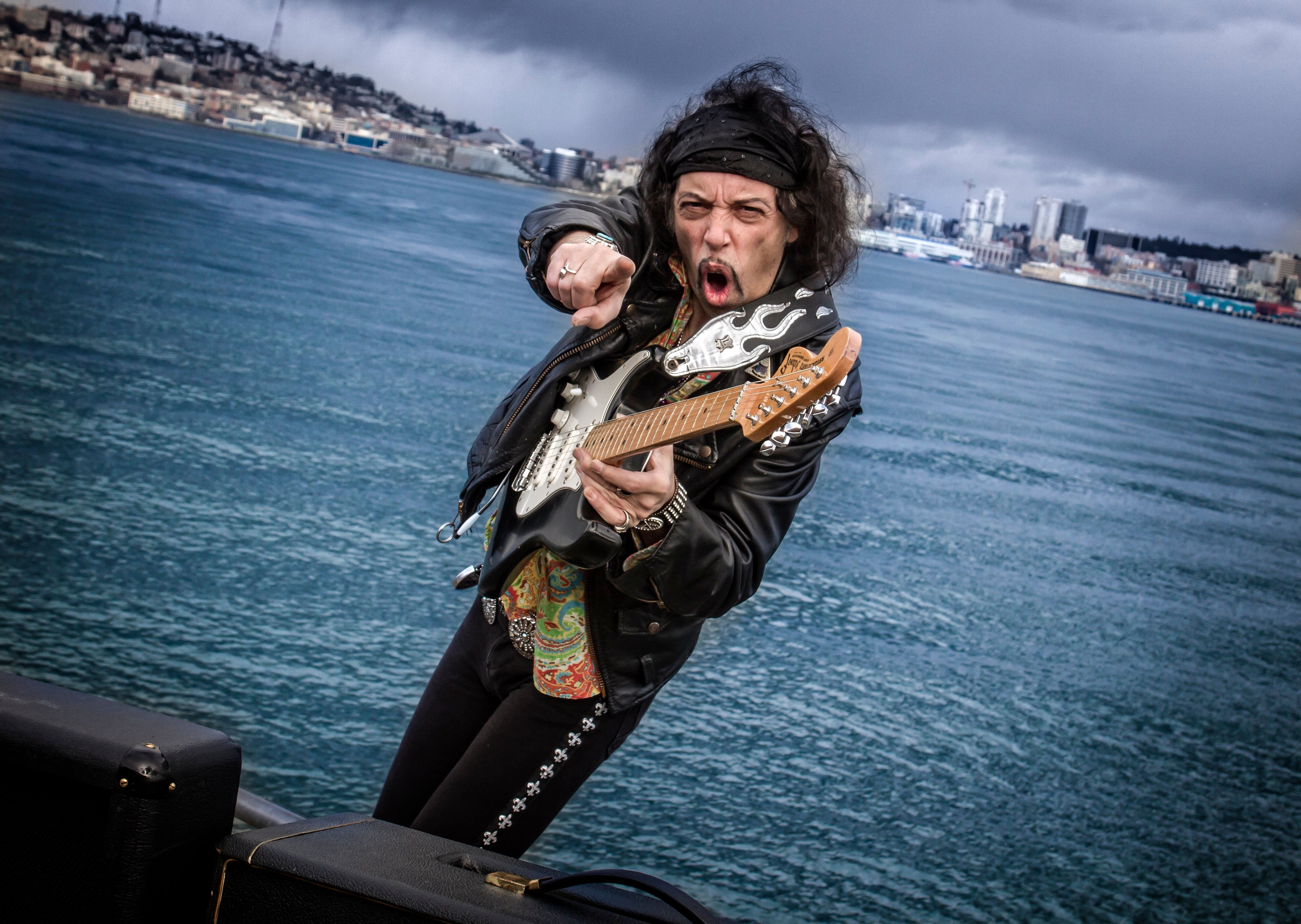 Ein Gitarrist in Lederjacke spielt leidenschaftlich E-Gitarre vor einer Küstenkulisse. Der Himmel ist bewölkt, und im Hintergrund sind Gebäude zu sehen.