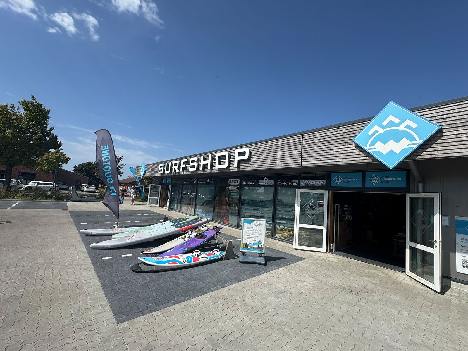 Ein Surfshop auf Fehmarn mit ausgestellten Surfbrettern und einer Fahne. Der Himmel ist klar und blau.