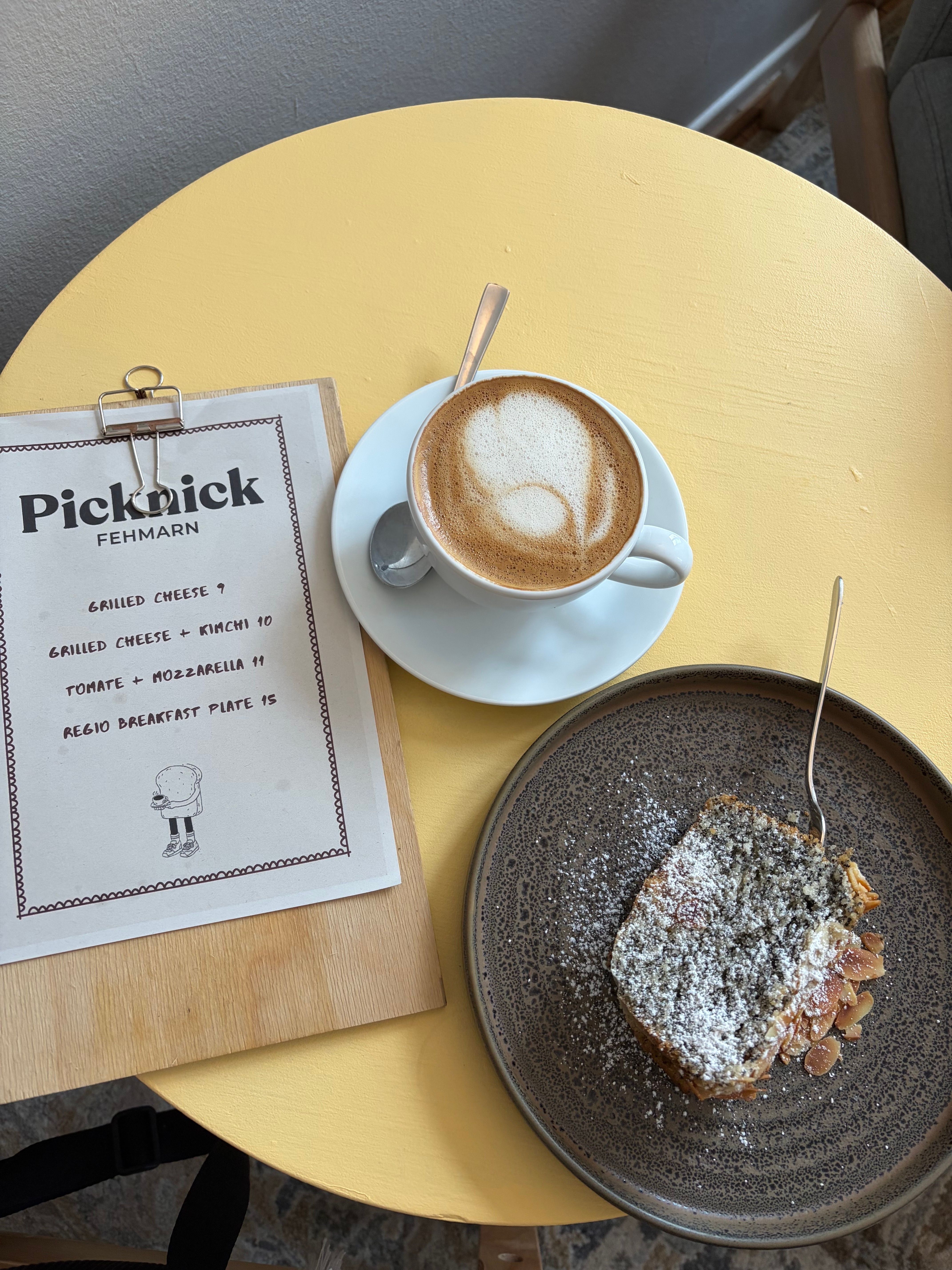 Auf einem gelben Tisch stehen eine Tasse Cappuccino, ein Stück Kuchen auf einem Teller und eine Speisekarte von Picknick Fehmarn.