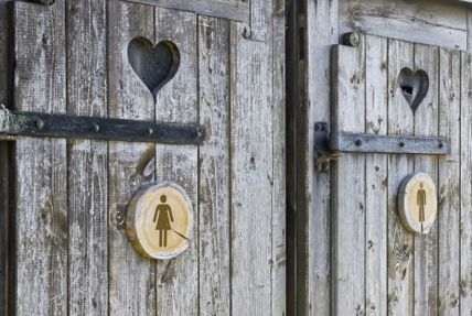 Zwei rustikale Holztüren mit Herzfenstern und Toilettensymbolen für Damen und Herren. Die Türen sind aus verwittertem Holz.