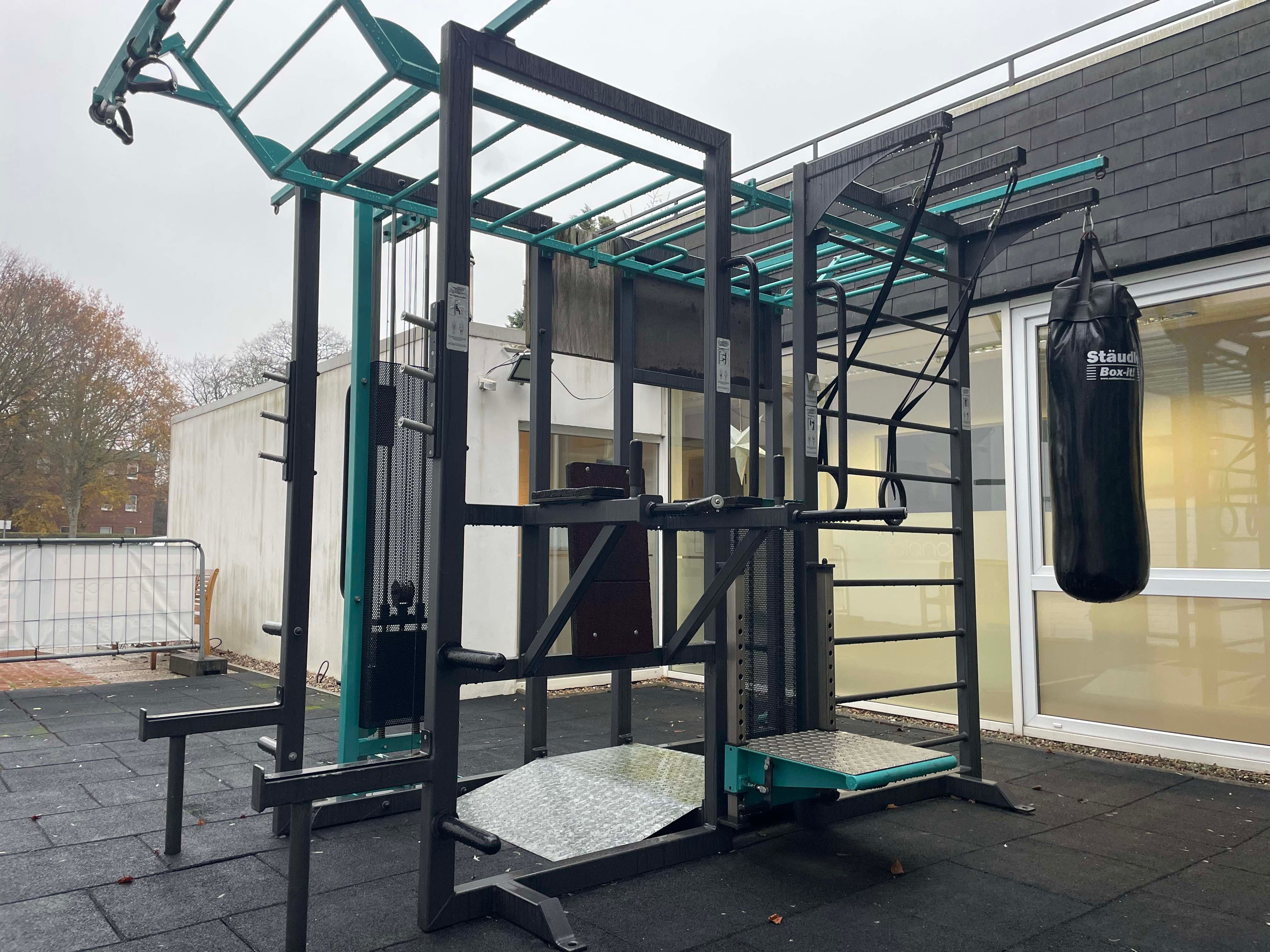 Ein Outdoor-Fitnessgerät mit Klettergerüst und Boxsack auf einem gepflasterten Boden vor einem Gebäude. Ideal für Kraft- und Ausdauertraining.