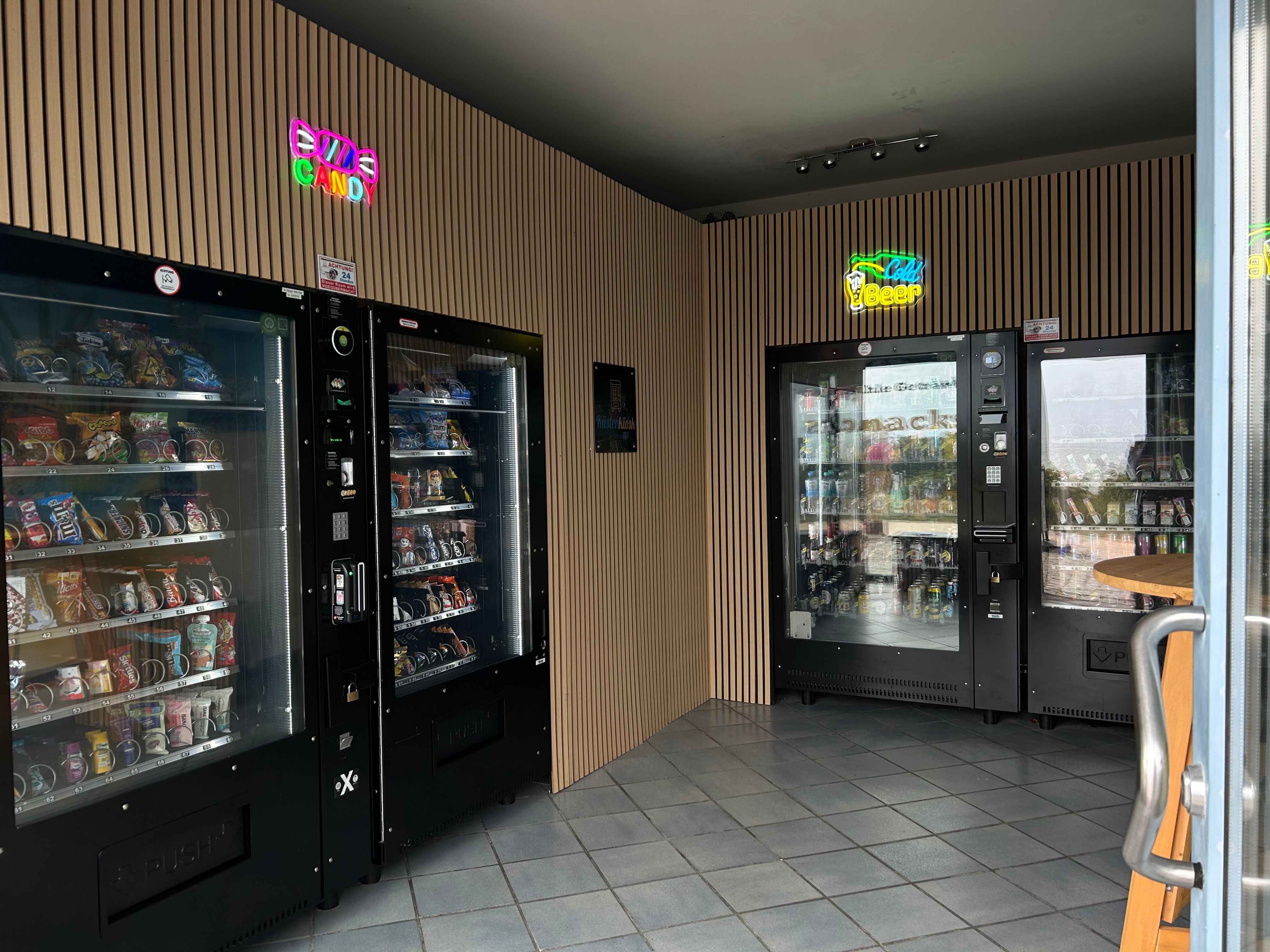 Ein kleiner Raum mit Snack- und Getränkeautomaten, beleuchtet von Neonlichtern mit den Aufschriften 'Candy' und 'Cold Beer'.