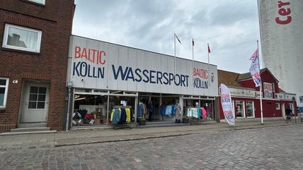 Ein Wassersportgeschäft namens 'Baltic Kölln' mit Schaufenstern voller Kleidung und Ausrüstung. Das Gebäude hat eine große Beschriftung und Flaggen.