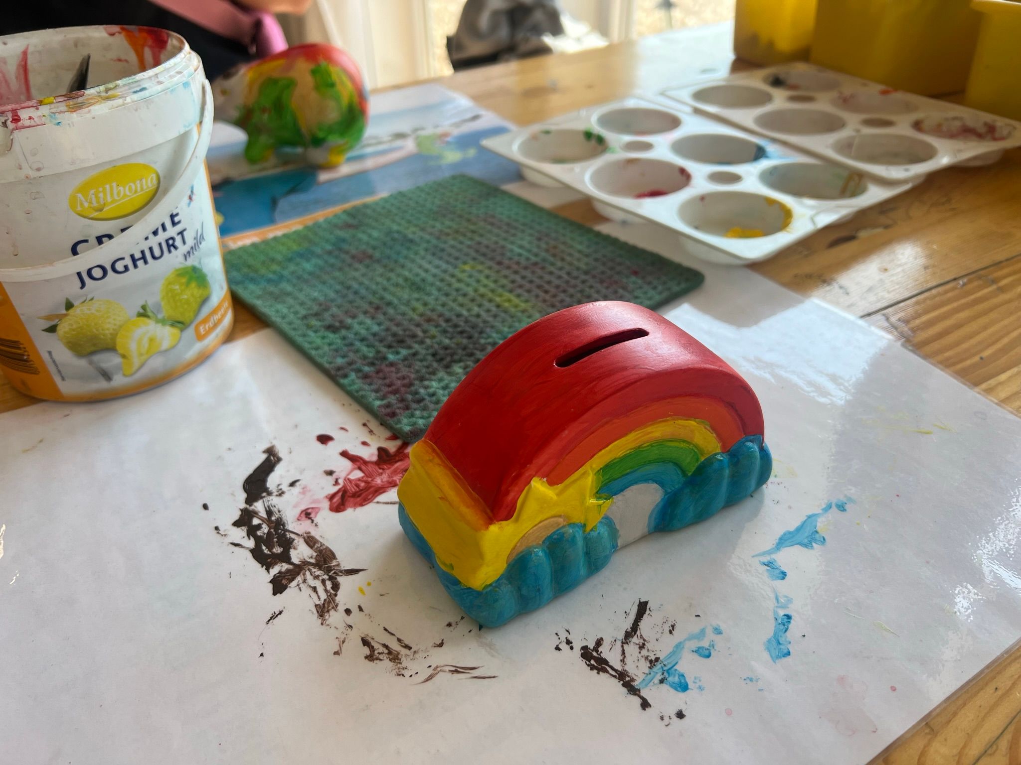 Ein bemalter Porzellan-Regenbogen auf einem Tisch mit Farbpaletten, einem Joghurtbecher und einem Schwamm.