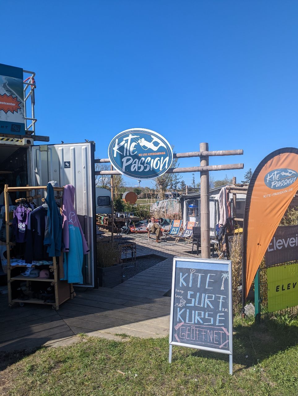 Eingang zur Kitesurf-Schule "Kite Passion" auf Fehmarn. Ein Schild weist auf geöffnete Surfkurse hin. Im Hintergrund sind Ausrüstungen und Kleidung zu sehen.