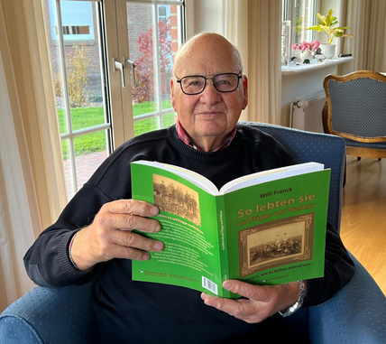 Ein älterer Mann sitzt in einem gemütlichen Sessel und liest ein Buch mit grünem Einband. Im Hintergrund ist ein großes Fenster mit Blick auf einen Garten.