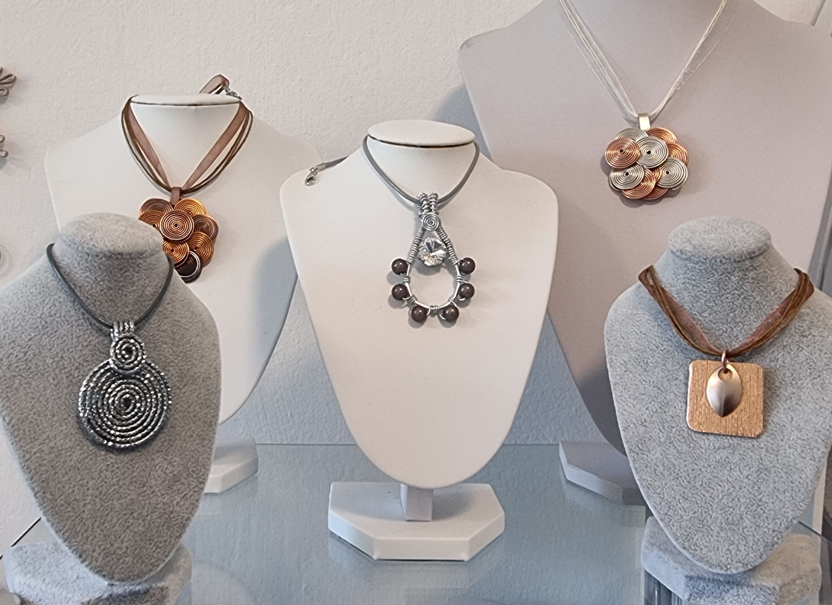 Eine Auswahl an kunstvollen Halsketten auf weißen und grauen Büsten präsentiert. Die Designs variieren von spiralförmigen Anhängern bis zu geometrischen Formen.