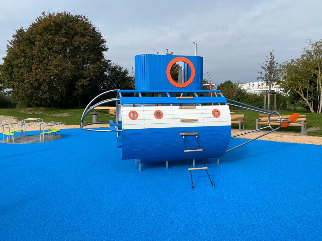 Ein Spielplatz mit einem blauen Spielschiff auf blauem Boden. Im Hintergrund sind Bäume und Bänke zu sehen.