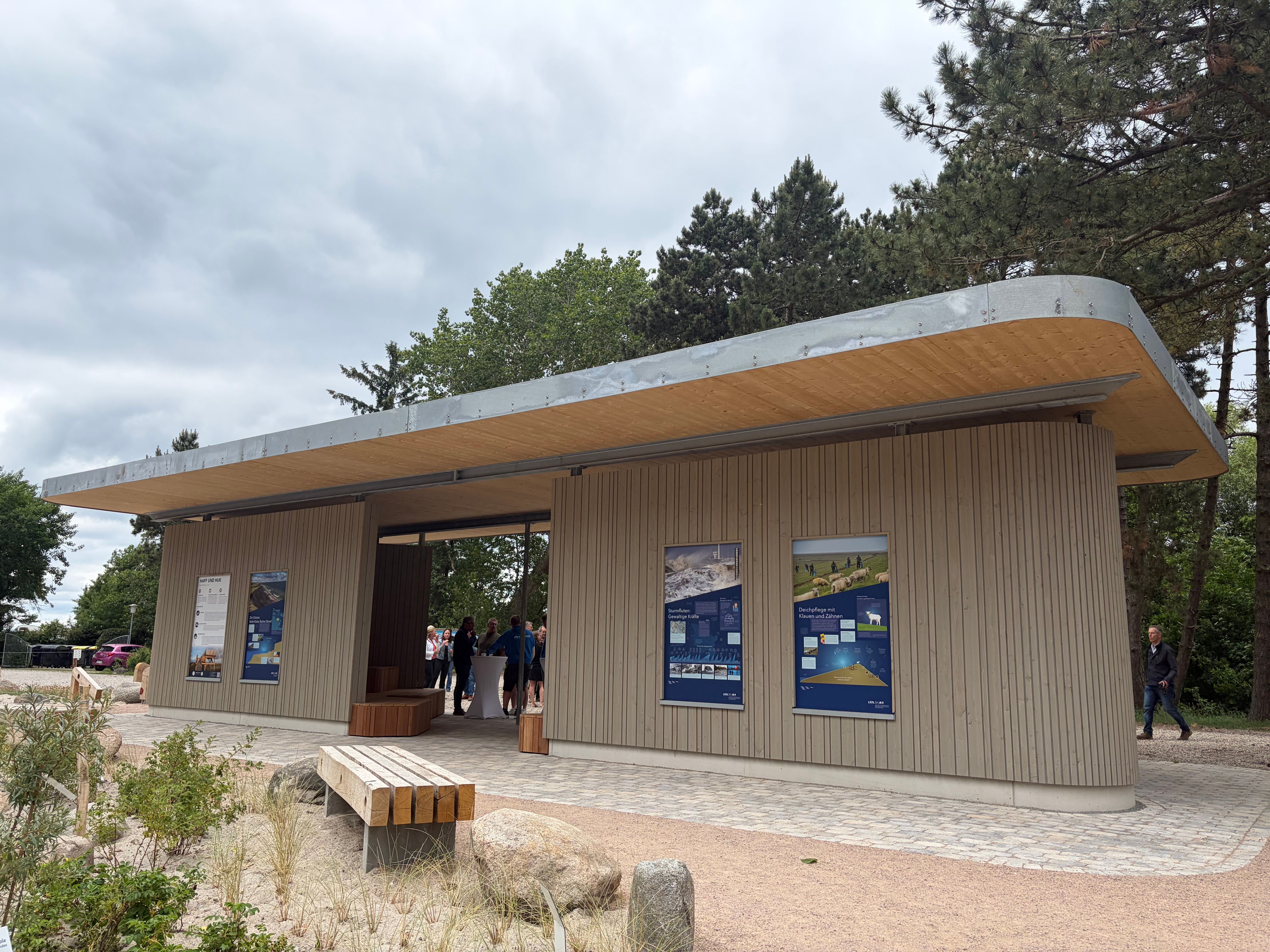 Moderner Info- und Schutzpavillon mit Holzverkleidung und Informationstafeln, umgeben von Bäumen und Pflanzen. Menschen stehen im Inneren.