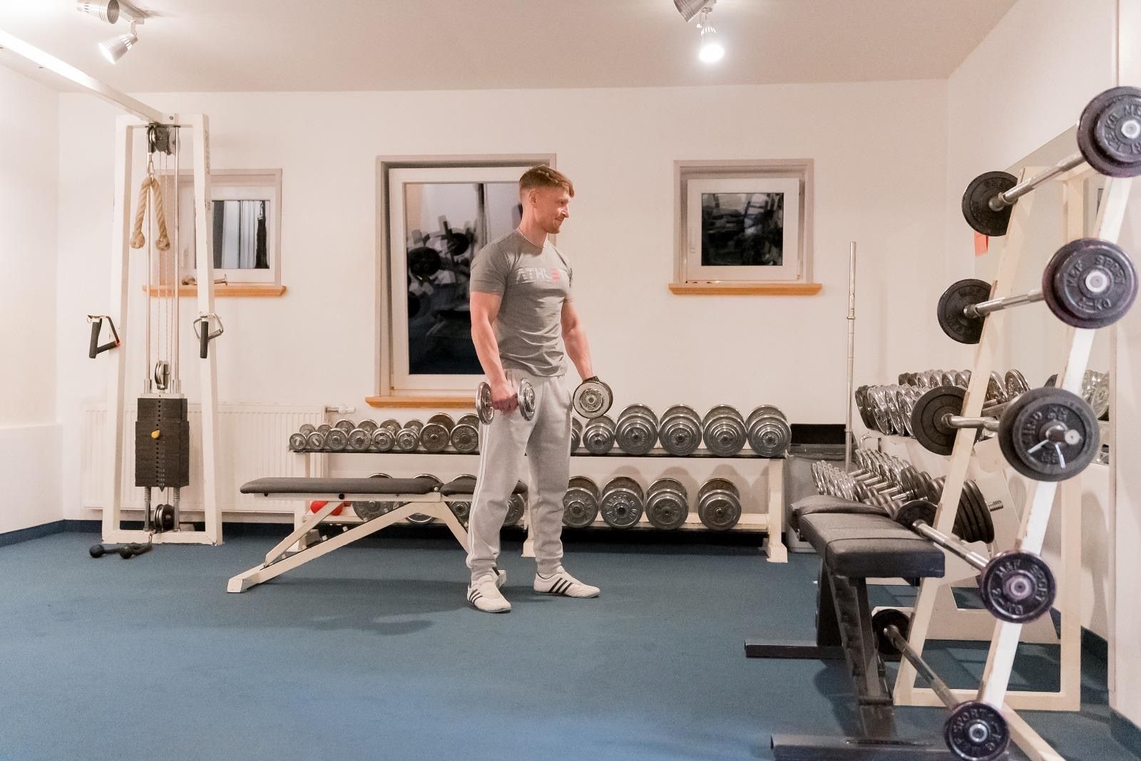 Ein Mann trainiert mit Hanteln in einem Fitnessstudio. Der Raum ist mit verschiedenen Gewichten und Fitnessgeräten ausgestattet.