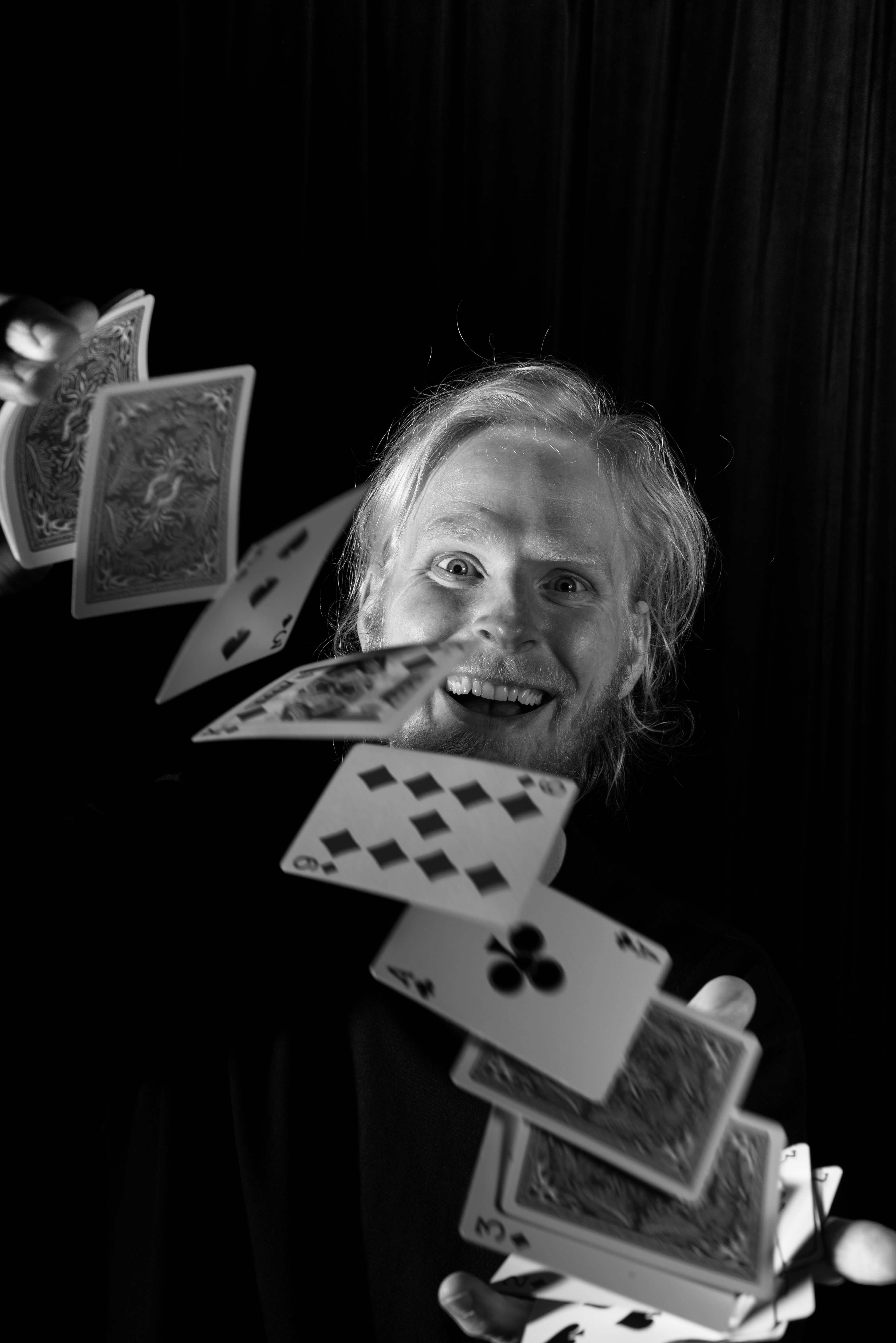 Eine Person mit langen Haaren wirft lächelnd Spielkarten in die Luft. Die Szene ist in Schwarz-Weiß gehalten.