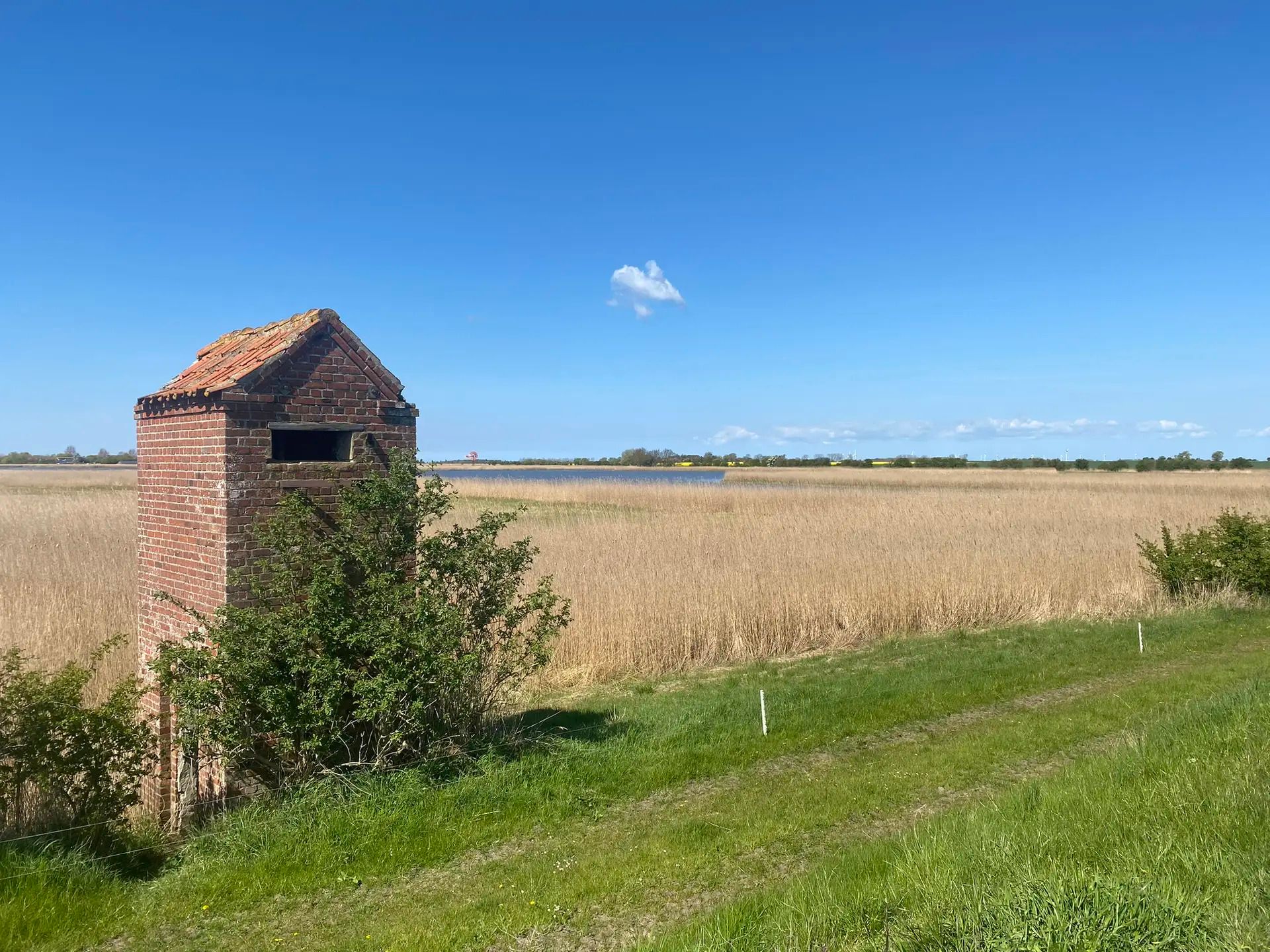 Ein altes Backsteingebäude steht in einer weiten Graslandschaft unter einem klaren blauen Himmel.