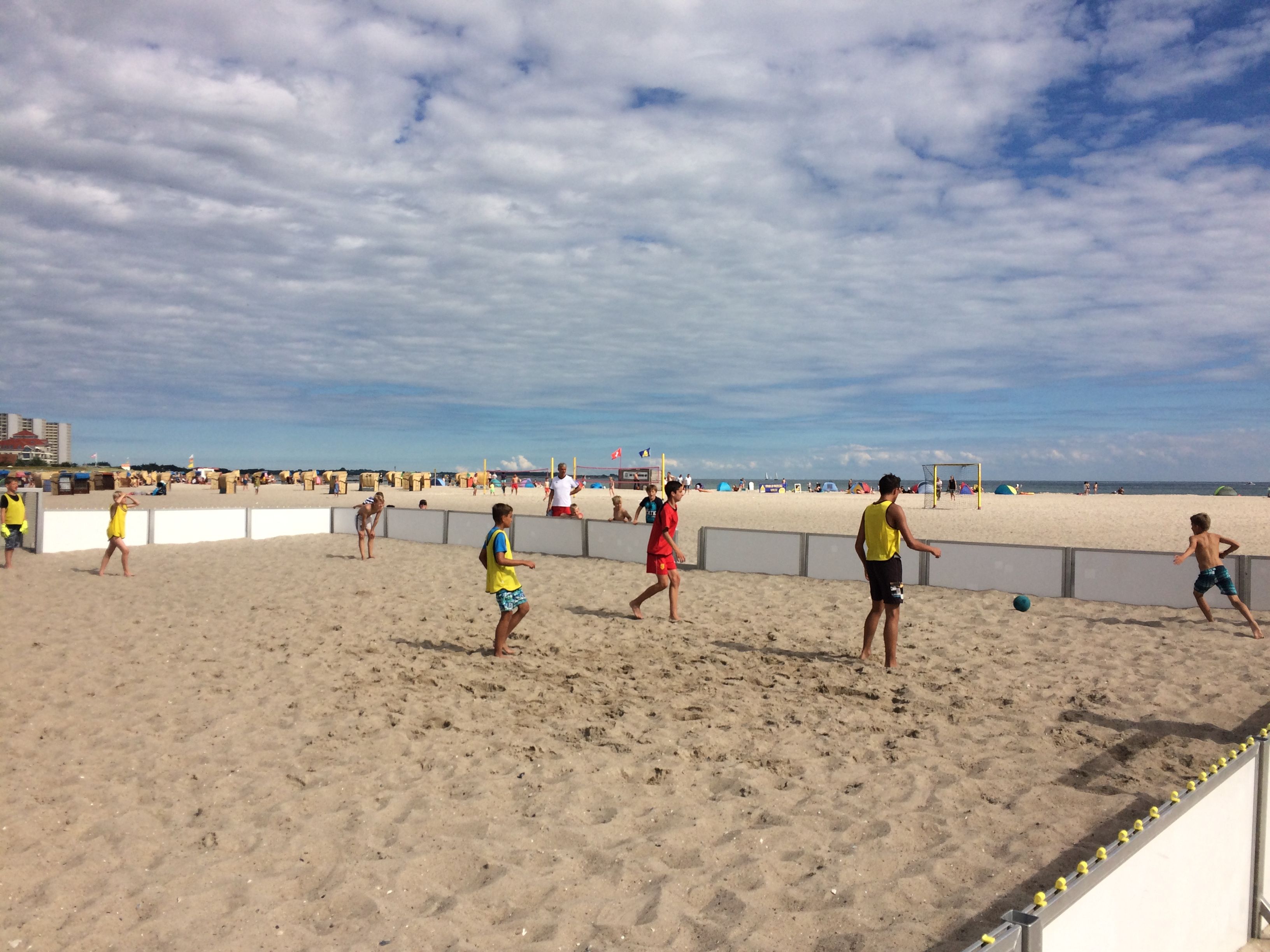 Jugendliche spielen Beachsoccer auf einem Sandplatz am Strand. Der Himmel ist bewölkt, und im Hintergrund sind Strandbesucher und das Meer zu sehen.