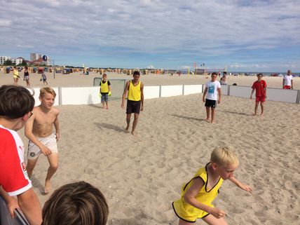 Kinder spielen Beachsoccer am Strand. Einige tragen gelbe Leibchen. Im Hintergrund sind Strandkörbe und ein bewölkter Himmel zu sehen.