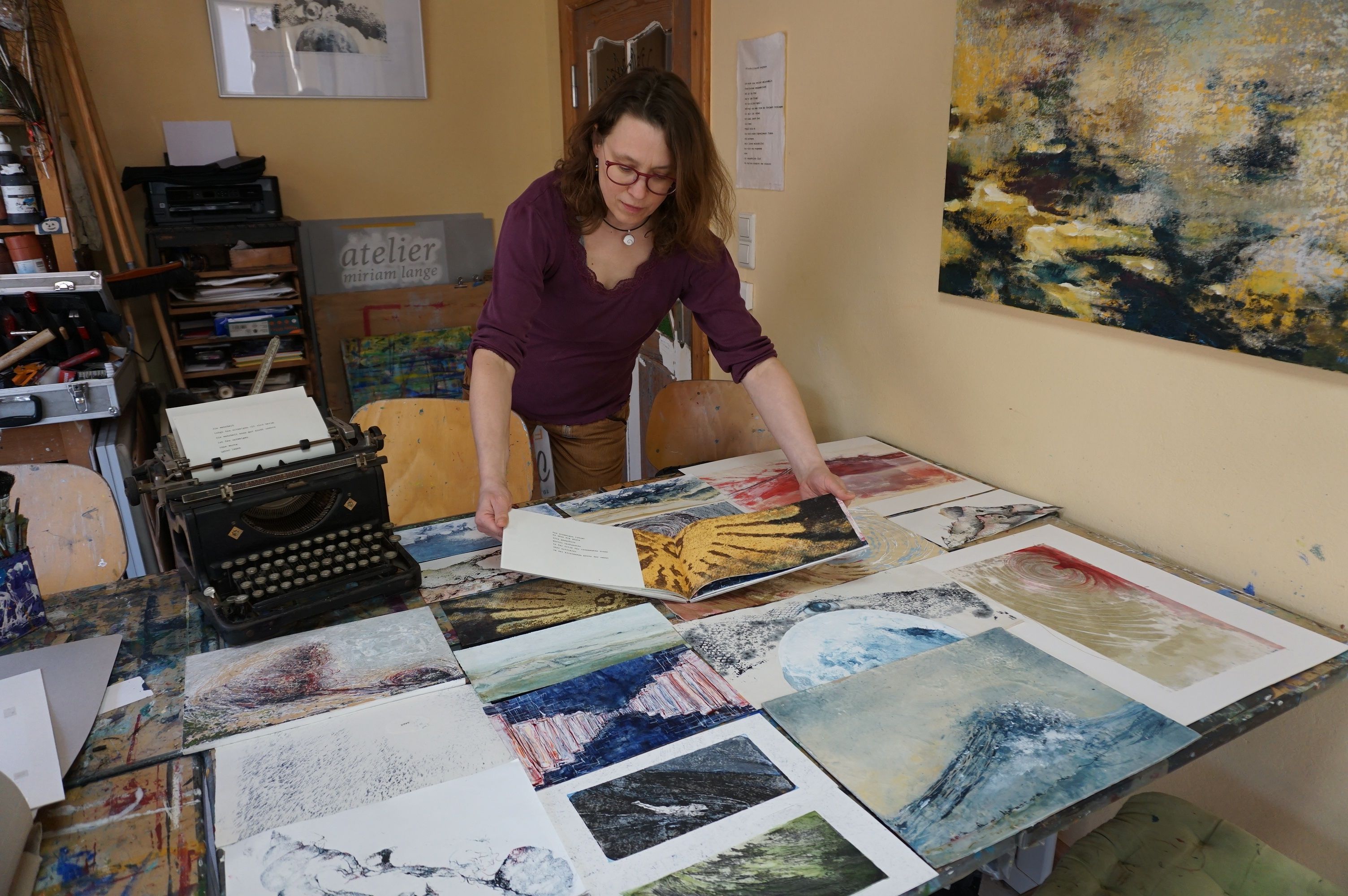 Eine Frau in einem Atelier betrachtet verschiedene Kunstwerke auf einem Tisch. Im Hintergrund sind eine Schreibmaschine und weitere Kunstmaterialien zu sehen.