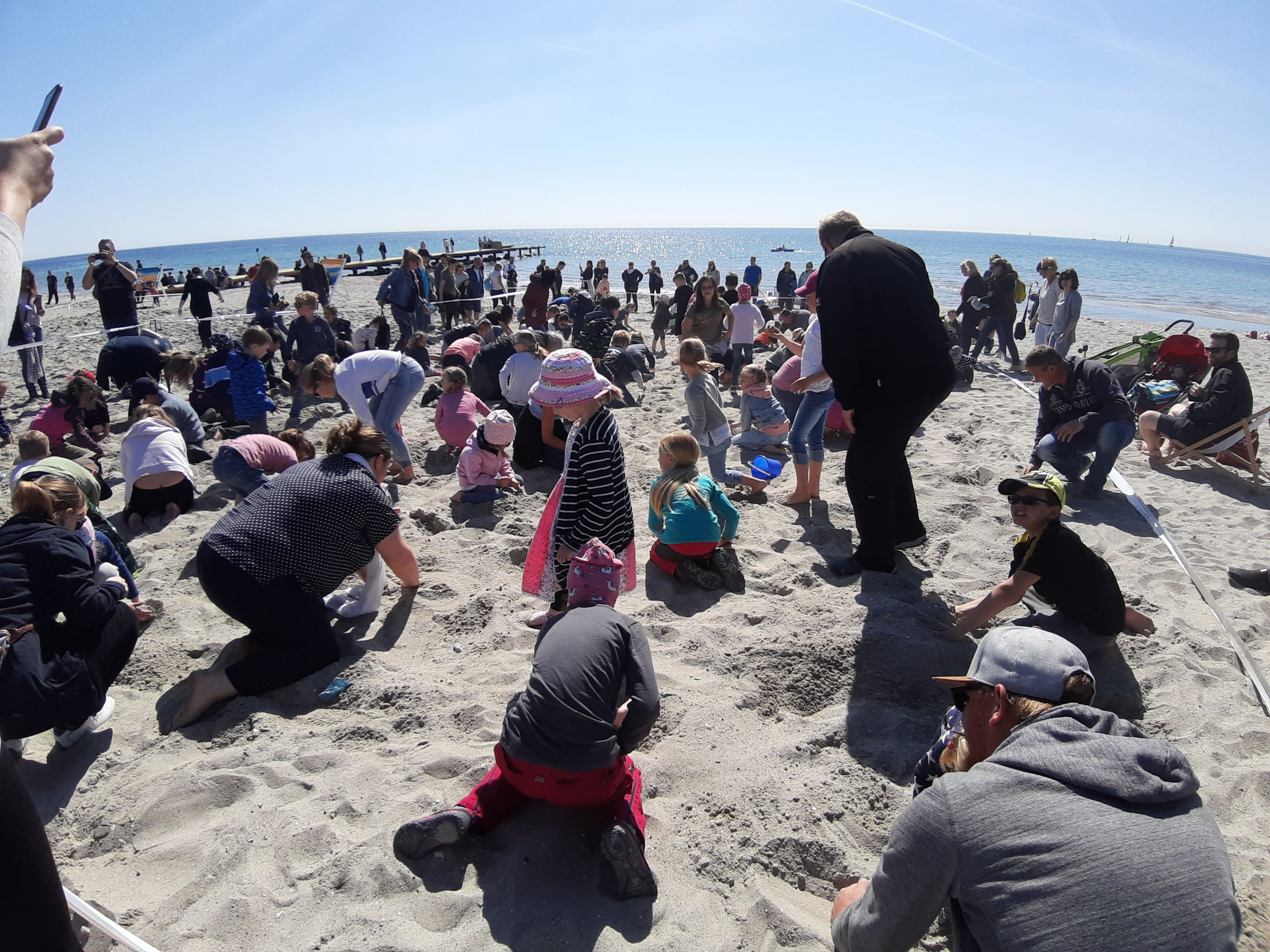 Viele Menschen, darunter Kinder und Erwachsene, graben am sonnigen Strand im Sand. Im Hintergrund ist das Meer zu sehen.