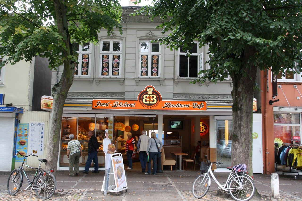 Eine Bäckerei mit Café in einer belebten Straße. Vor dem Geschäft stehen Fahrräder, und Passanten schauen in die Auslage.
