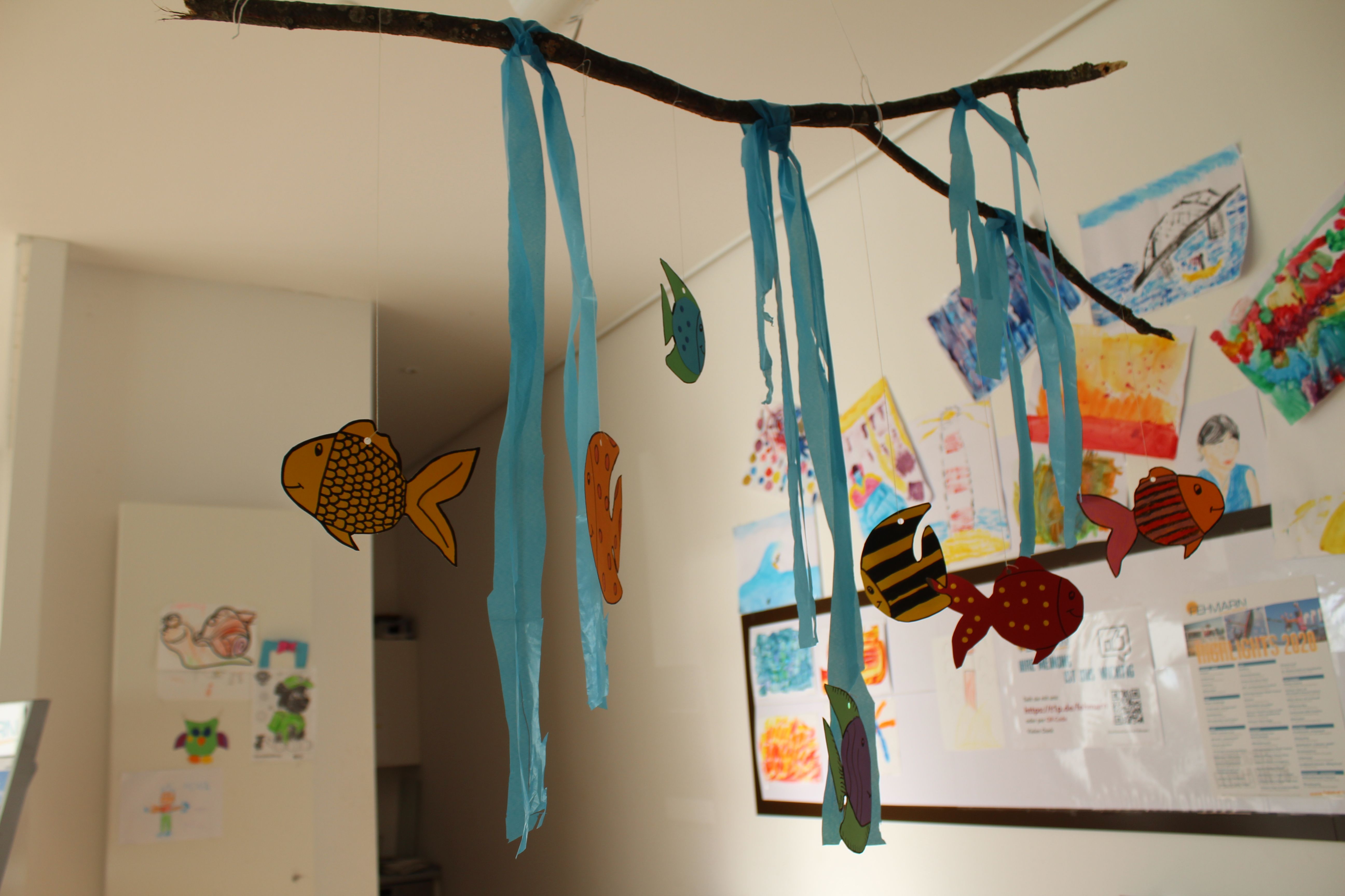 Ein Mobile aus bunten Papierfischen hängt an einem Ast mit blauen Bändern. Im Hintergrund sind Kinderzeichnungen an der Wand.