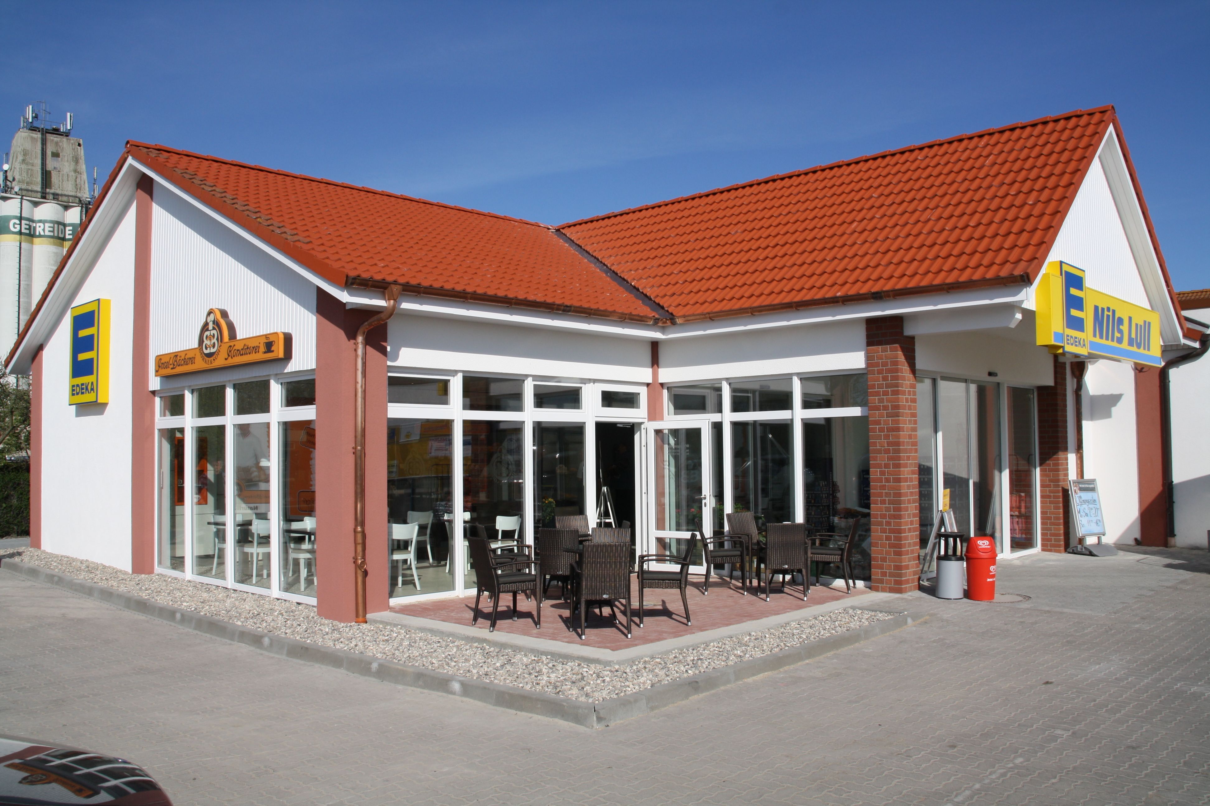 Ein Edeka-Supermarkt mit rotem Ziegeldach und angrenzendem Café. Vor dem Gebäude stehen Tische und Stühle für Gäste.
