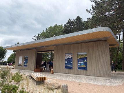 Moderner Info- und Schutzpavillon mit Holzverkleidung und Informationstafeln, umgeben von Bäumen und Pflanzen. Menschen stehen im Inneren.