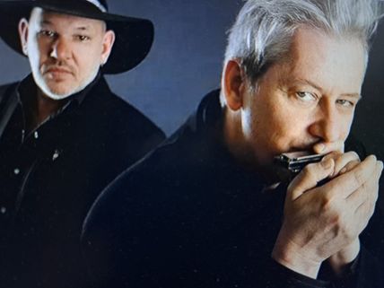 Zwei Männer in dunkler Kleidung, einer spielt Mundharmonika, der andere trägt einen Hut und schaut in die Kamera.