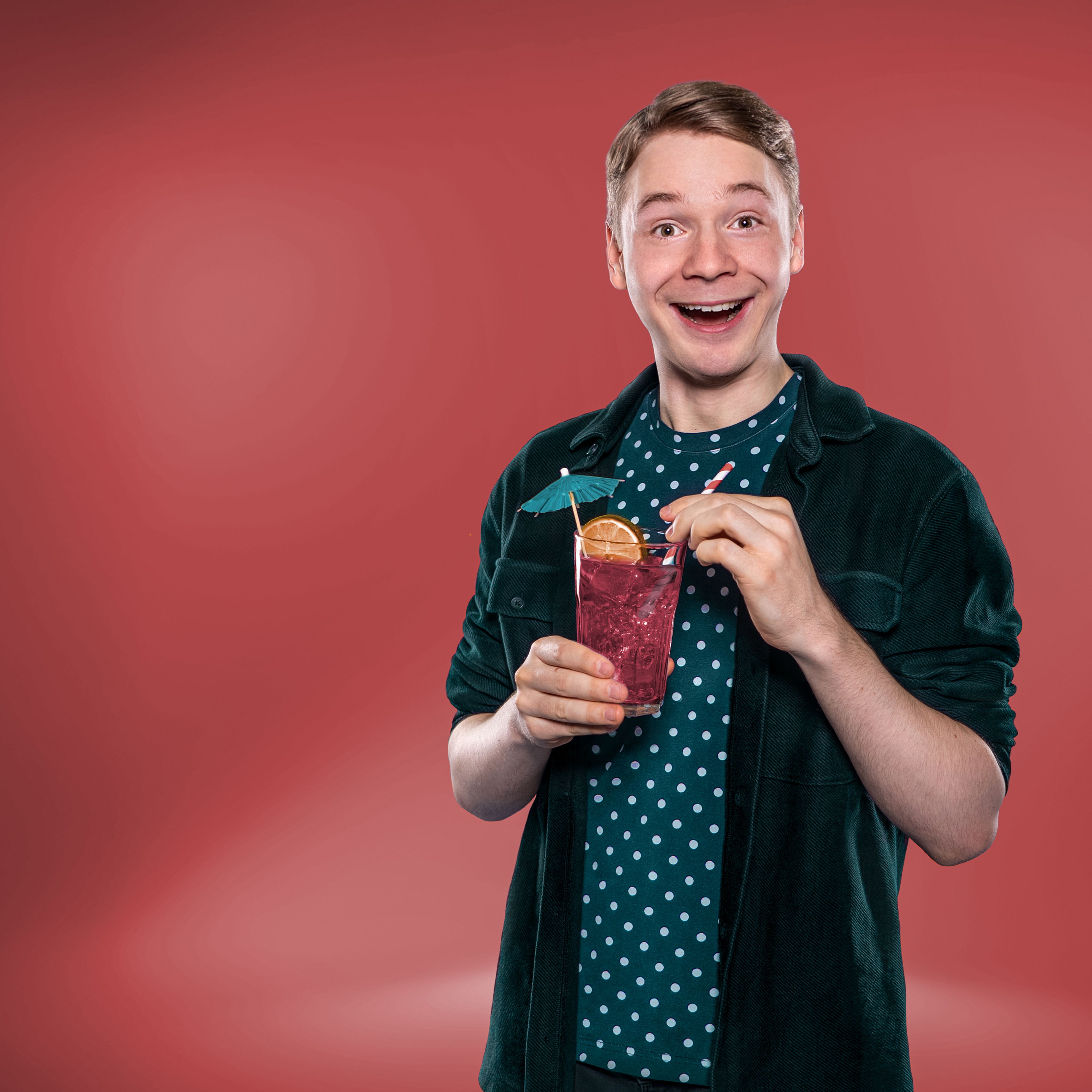 Ein fröhlicher Mann mit einem Getränk in der Hand, dekoriert mit einem Schirm und einer Orangenscheibe, vor einem roten Hintergrund.