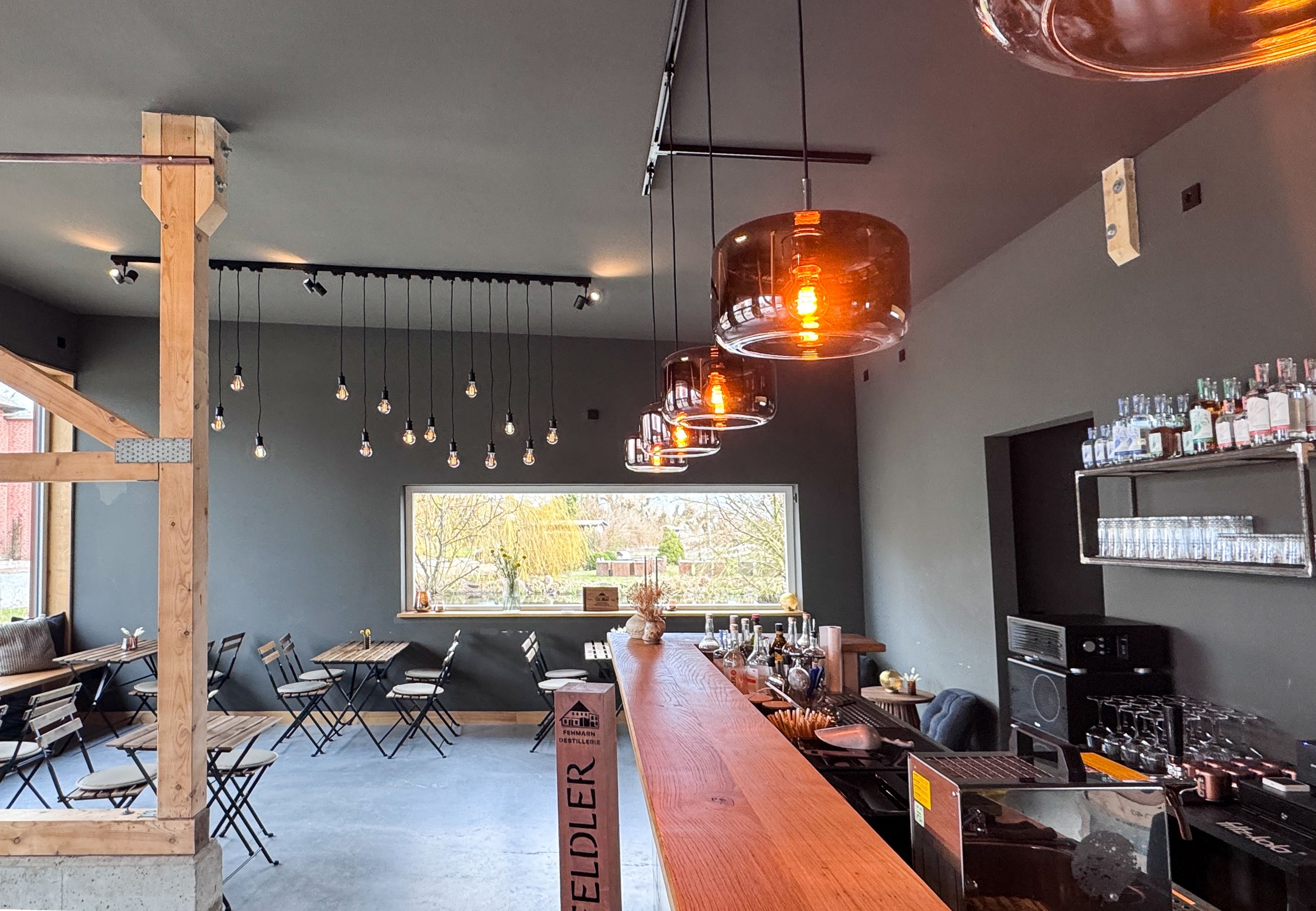Moderne Bar mit hölzernem Tresen, hängenden Glühbirnen und gemütlichen Sitzgelegenheiten. Große Fenster bieten Blick nach draußen.