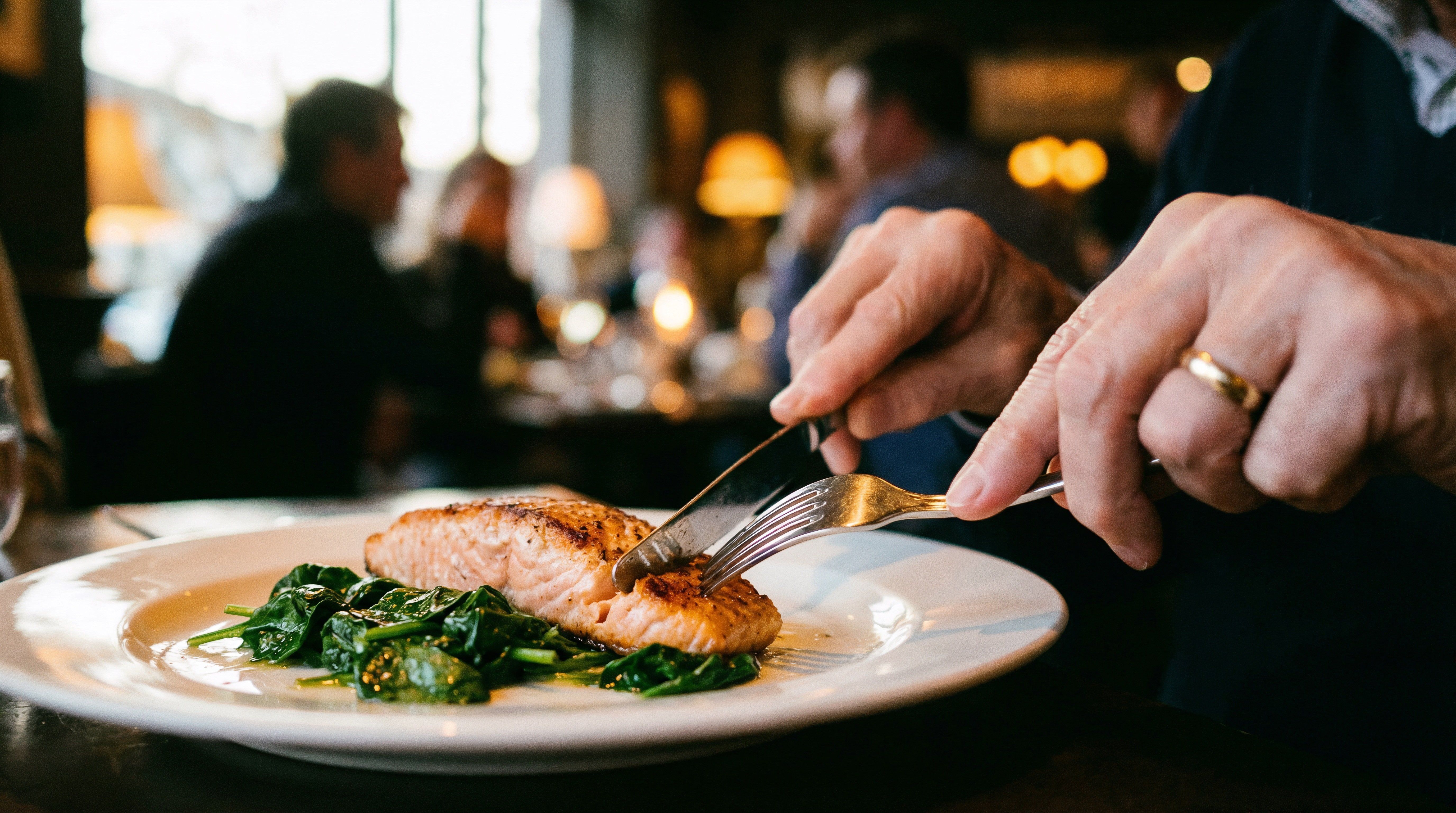 Ein Teller mit gebratenem Lachs und Spinat wird in einem gemütlichen Restaurant serviert. Eine Person schneidet den Lachs mit Messer und Gabel.