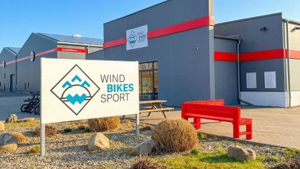 Ein modernes Gebäude mit dem Schild 'Wind Bikes Sport' und einer roten Bank im Vordergrund. Fahrräder stehen an der Seite bereit.