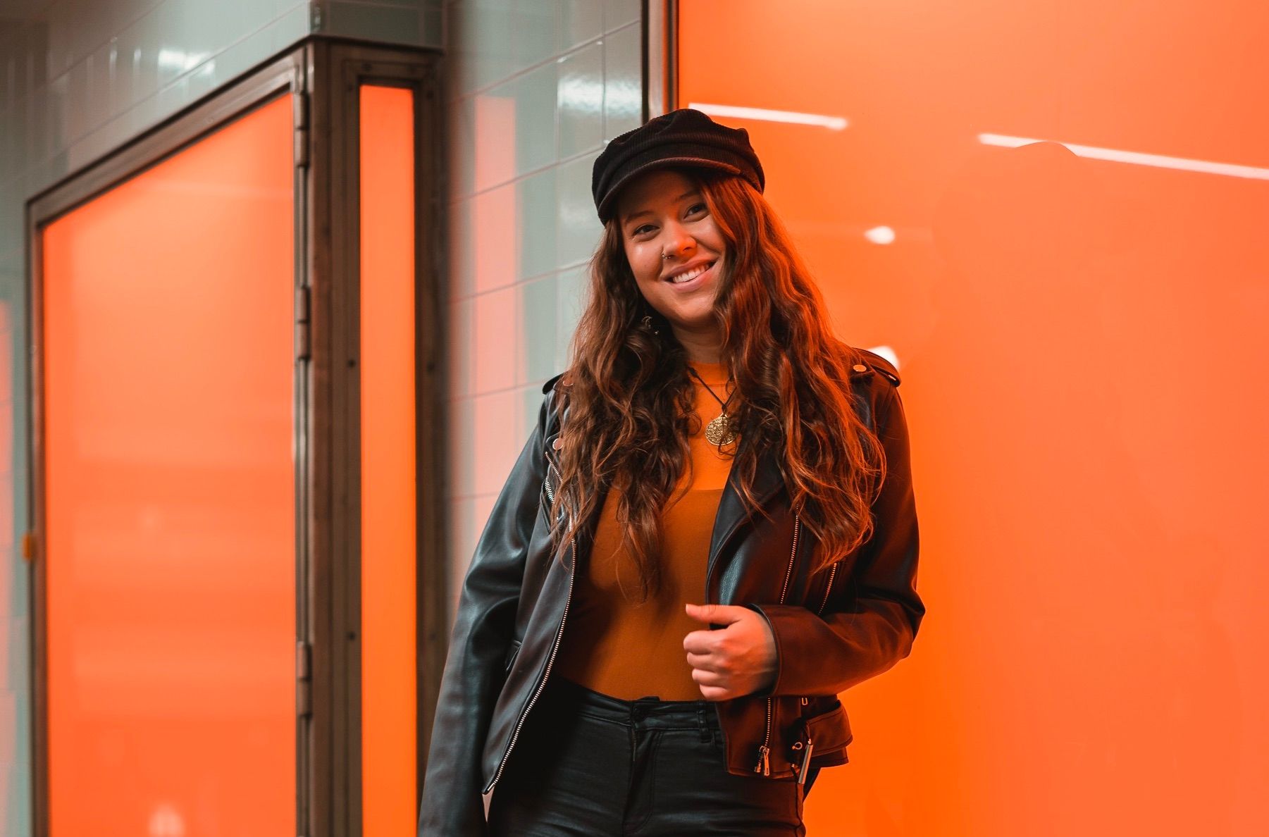 Eine Frau mit langen Haaren trägt eine schwarze Lederjacke und eine Mütze. Sie steht vor einem orange beleuchteten Hintergrund und lächelt in die Kamera.
