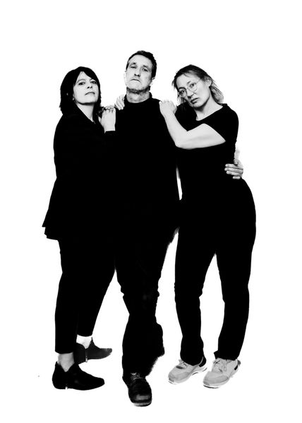 Drei Personen in Schwarz-Weiß posieren vor einem weißen Hintergrund. Sie stehen eng beieinander und tragen dunkle Kleidung.