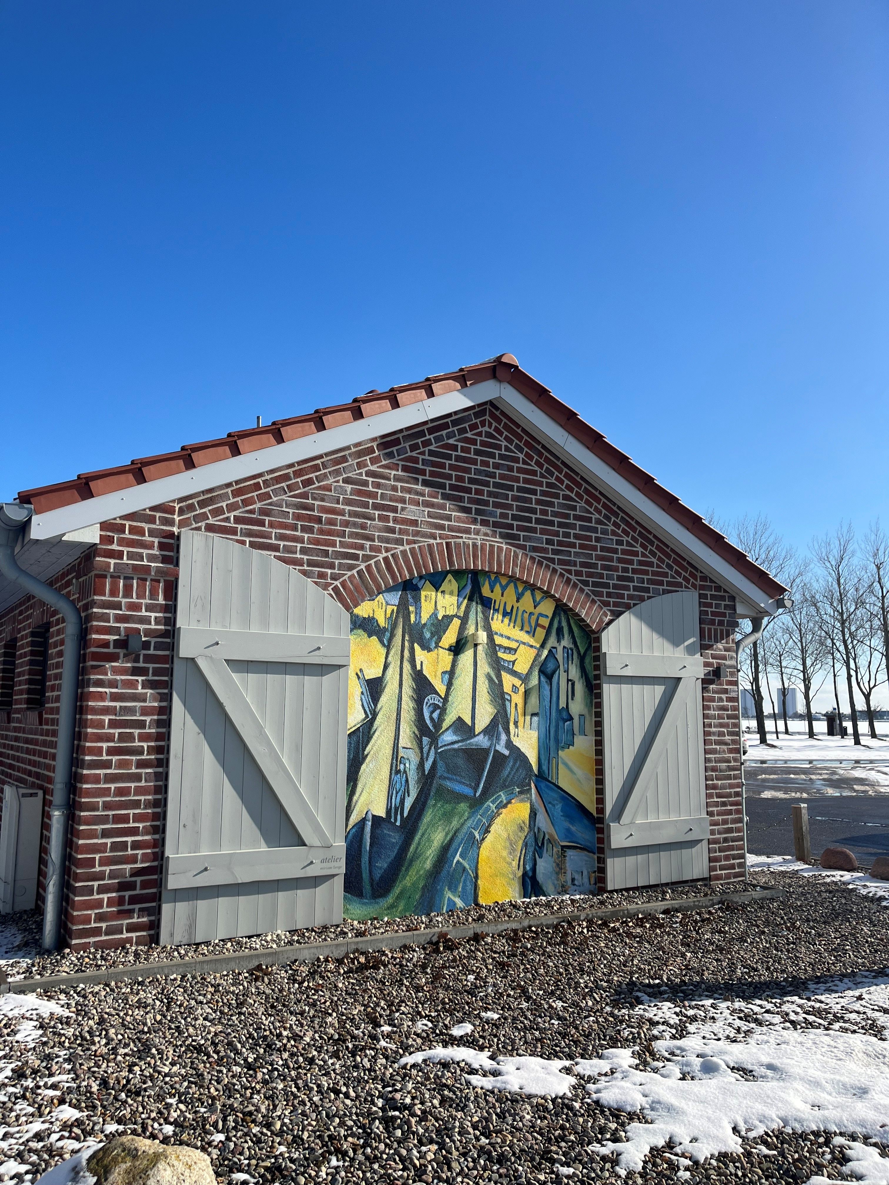 Ein Backsteingebäude mit einem farbenfrohen Wandgemälde von Segelbooten. Der Boden ist mit Kies und Schnee bedeckt, der Himmel ist klar und blau.